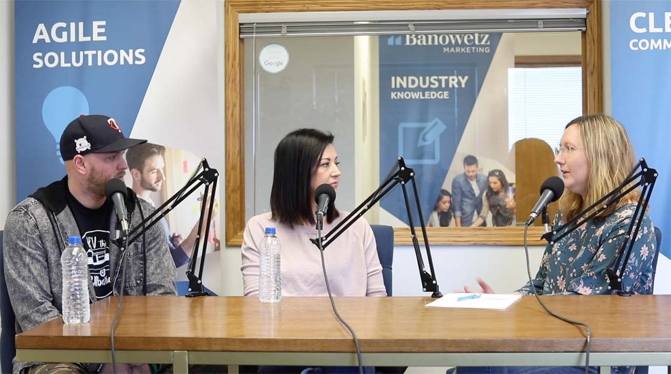Man and woman sitting at a table in front of microphones talking to a woman at the other end of the table while they record a podcast.