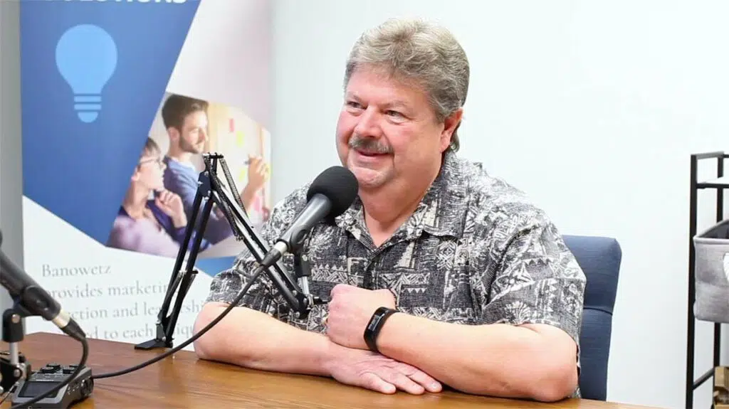 Man smiling and talking while sitting at a table in front of a microphone as a podcast is recorded.