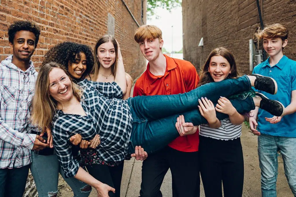 6 teenagers are standing in a line holding their mom who is laughing in a family photo. There are two brick walls in the background.