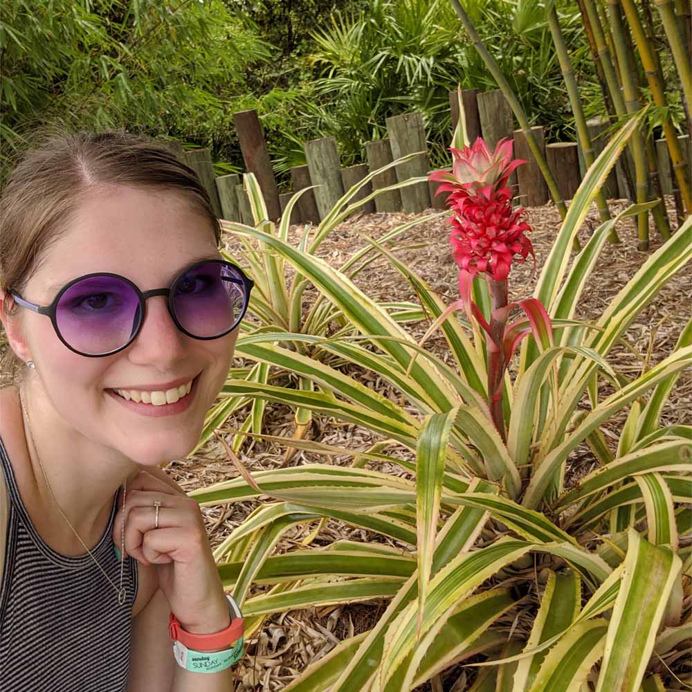 Woman posing in front of a tiny pink pineapple in a garden.